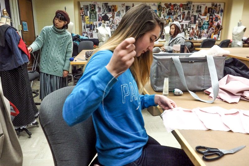 Student sewing on a table with other students behind working on a dress form.